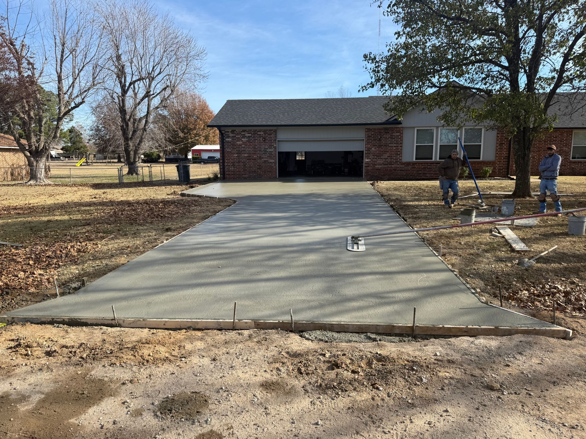 Freshly poured concrete driveway in front of a brick house; two workers smoothing the surface.