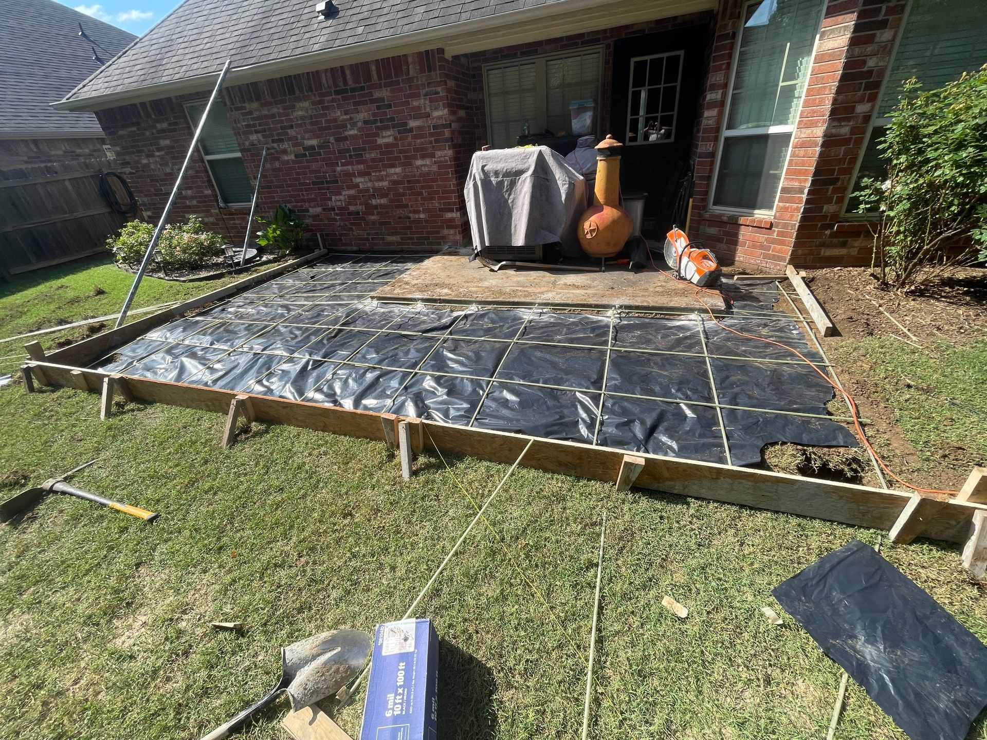 Backyard patio under construction with black plastic sheeting and wooden framing beside a brick house