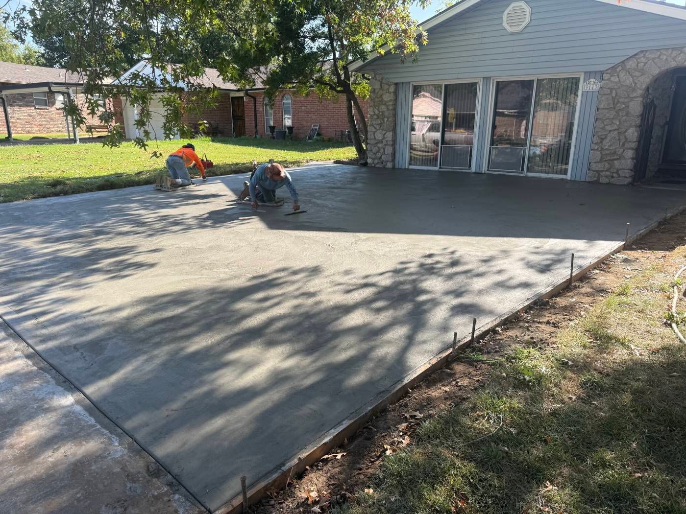 Two workers smoothing a fresh concrete patio beside a house in a sunny backyard