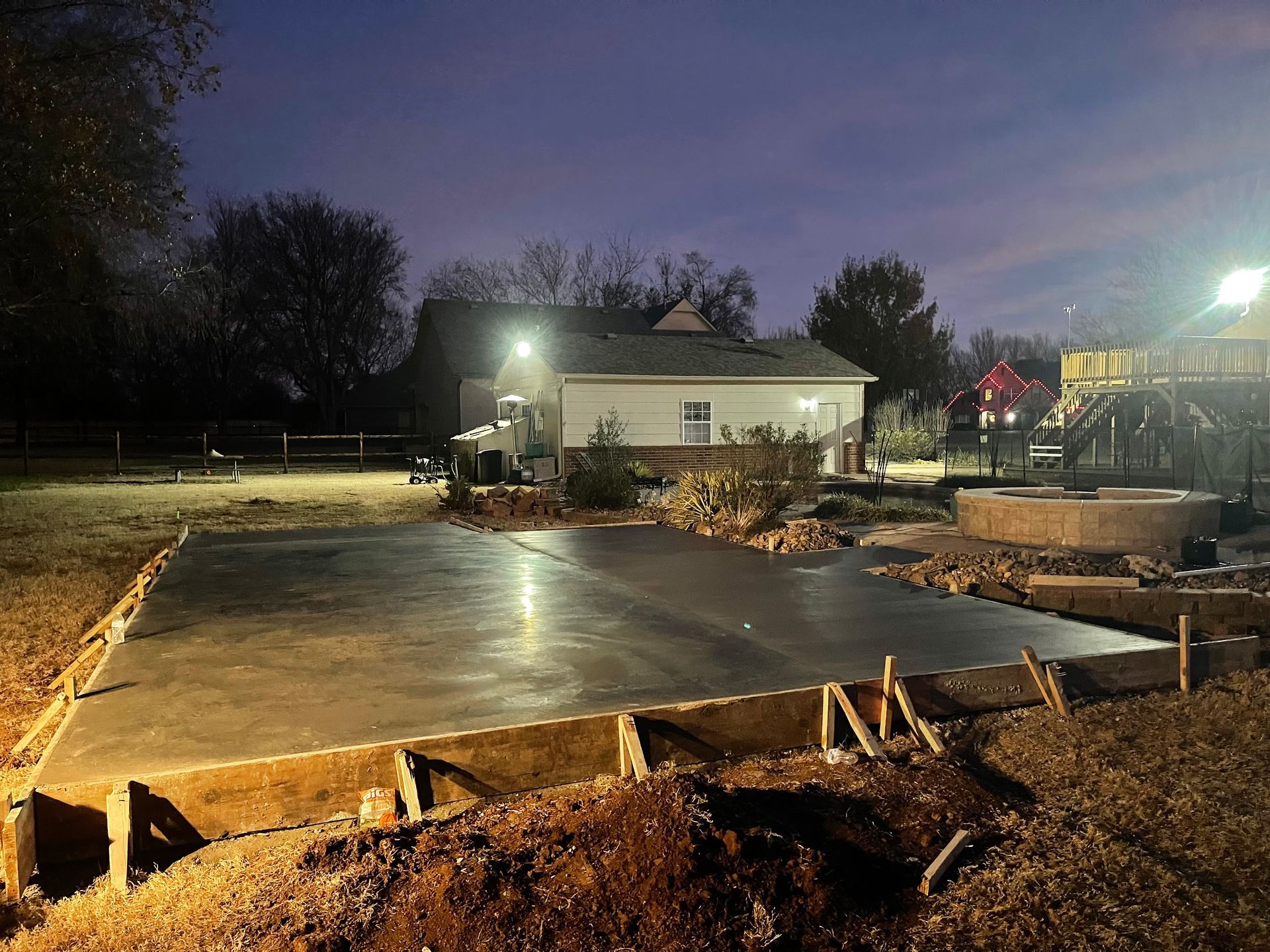 Newly poured concrete slab in a yard, framed by wooden forms, lit by outdoor lights, house in background.