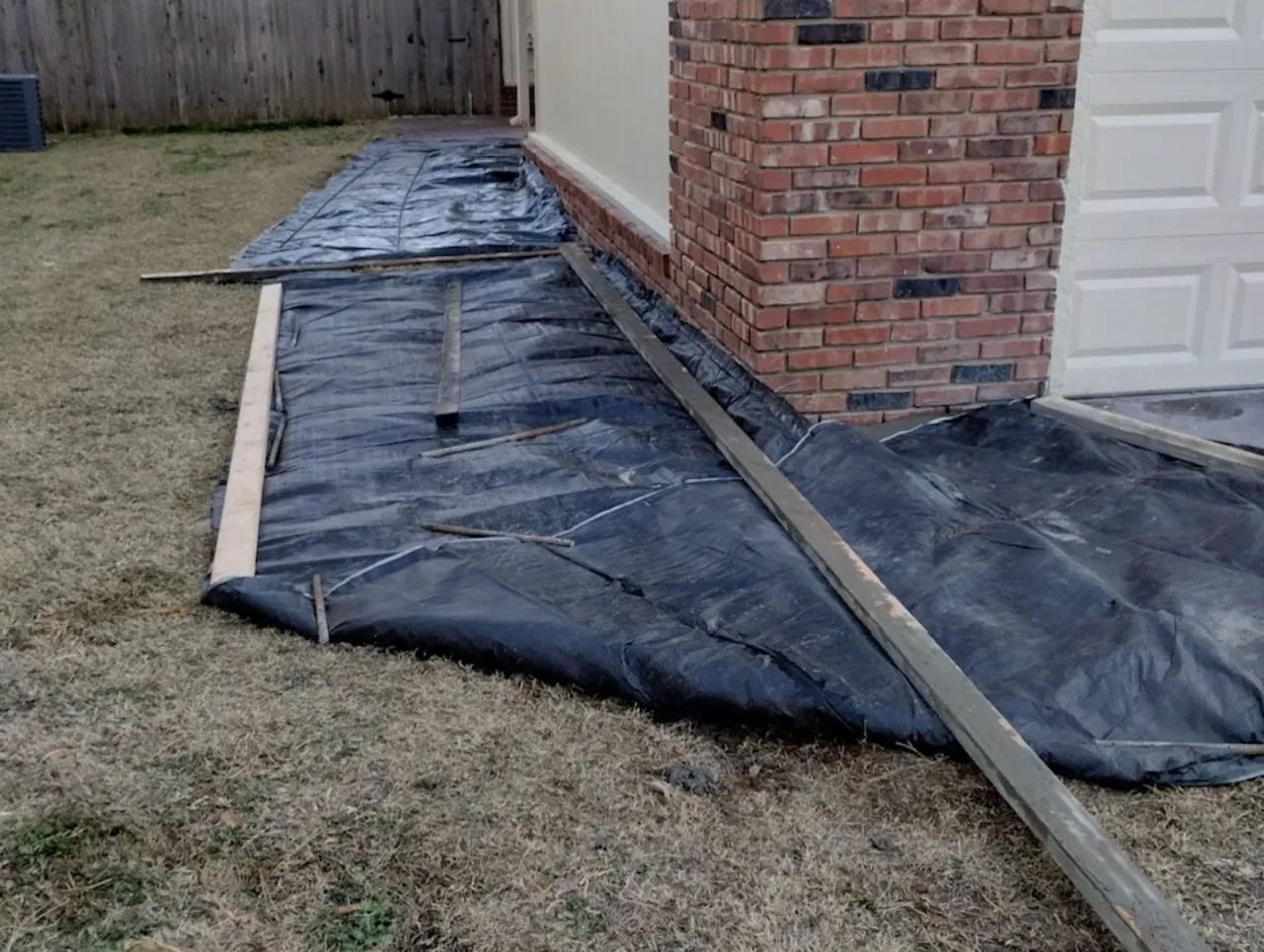Black landscape fabric laid on grass next to a brick wall and garage door. Wooden edging surrounds the fabric.