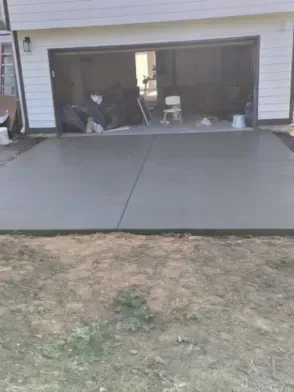 New concrete driveway in front of a garage; gray surface with dark border, surrounded by dirt and grass.