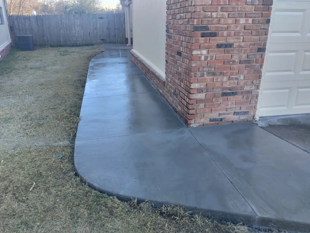 A newly poured concrete sidewalk curves alongside a brick and cream-colored wall; it is wet.