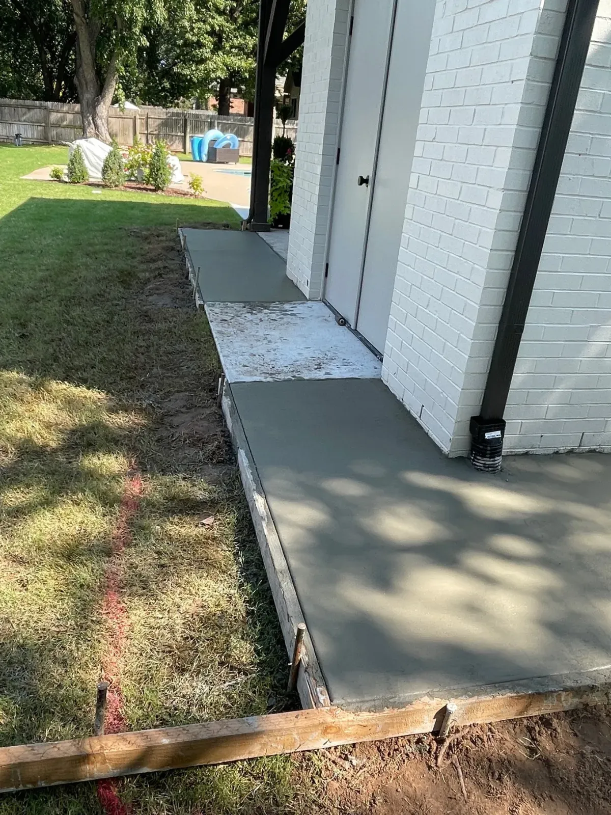 A concrete walkway next to a white brick building. Green grass and a dark rain gutter are visible.