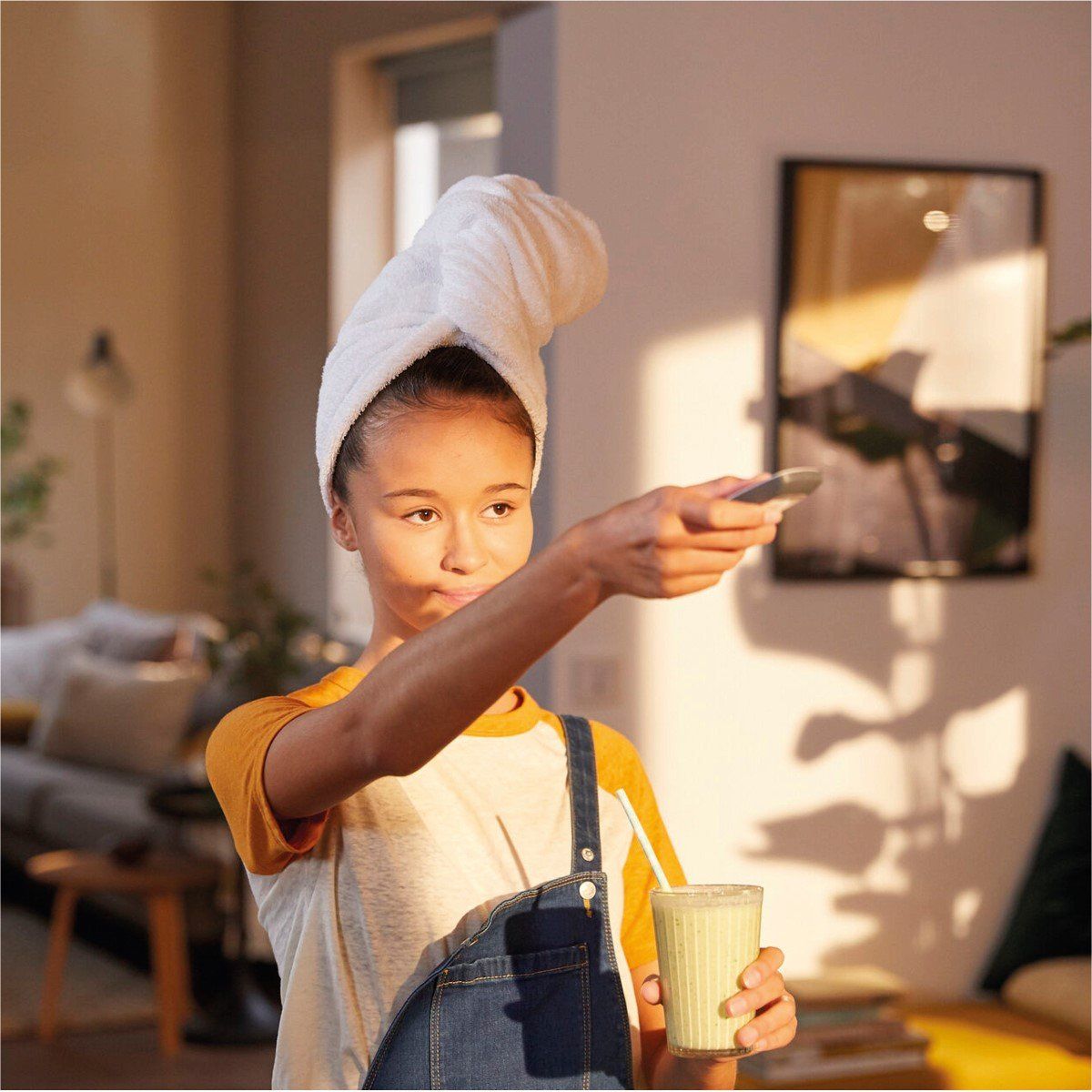 A young girl with a towel wrapped around her head is holding a cup and a cell phone.