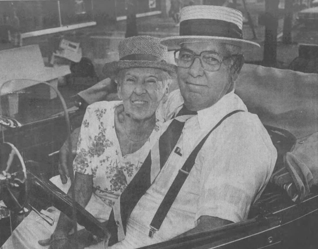 Elderly couple in vintage car, smiling. Man wears straw hat, suspenders. Woman in floral top.