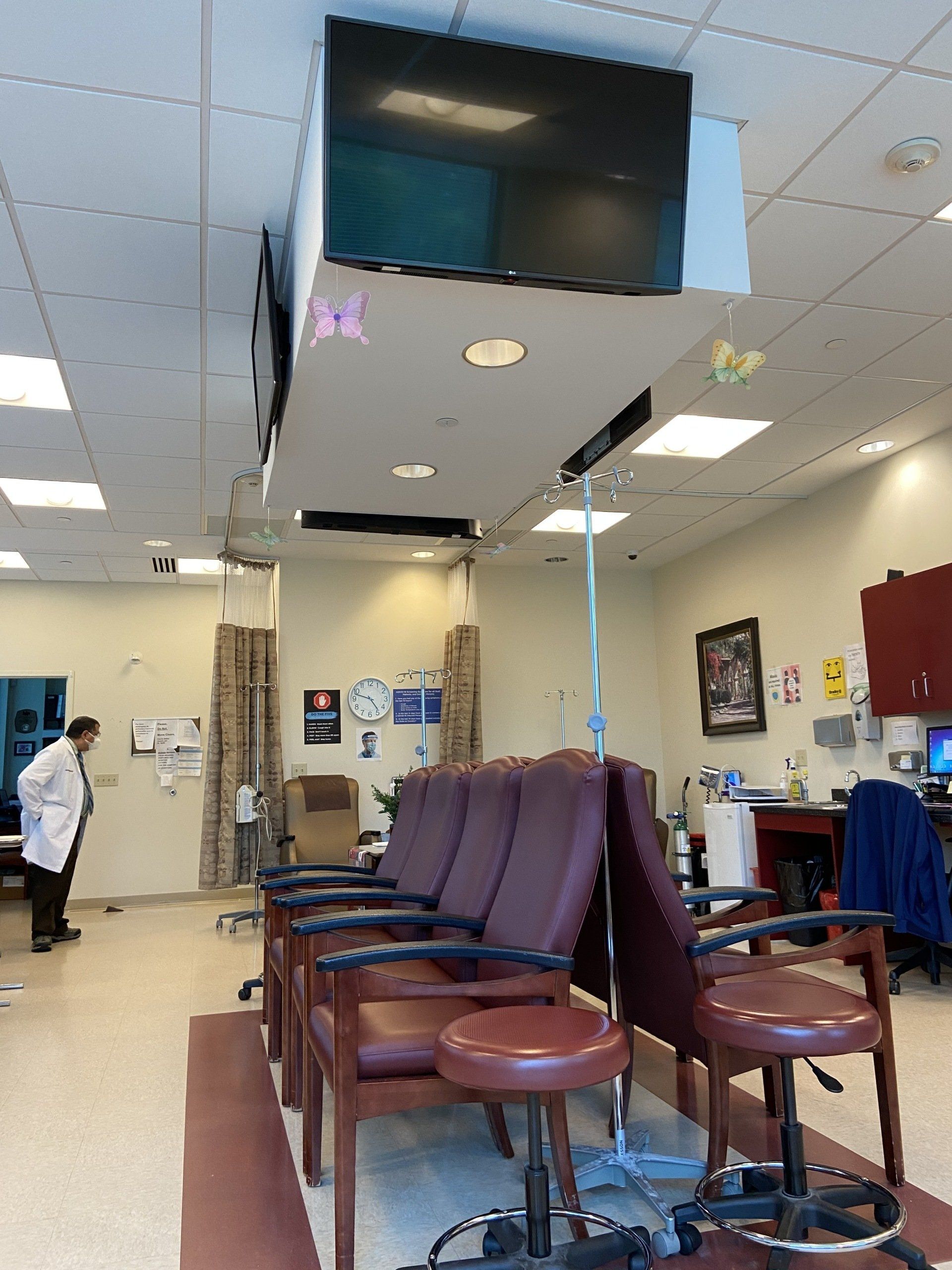 Medical infusion room with chairs, IV pole, and ceiling-mounted televisions. A person in a white coat stands in the background.