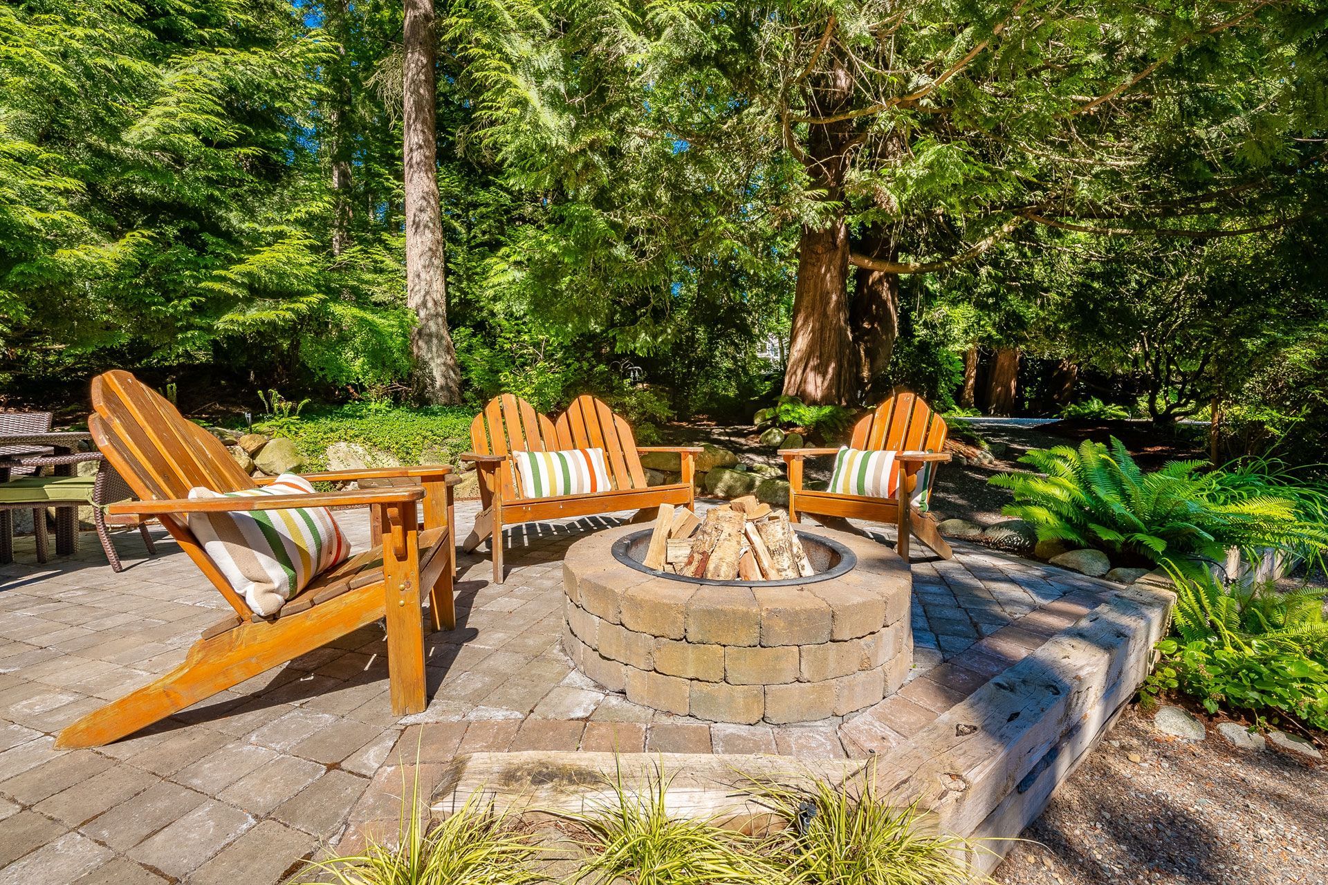Wooden Adirondack chairs around a stone fire pit in a sunny wooded backyard