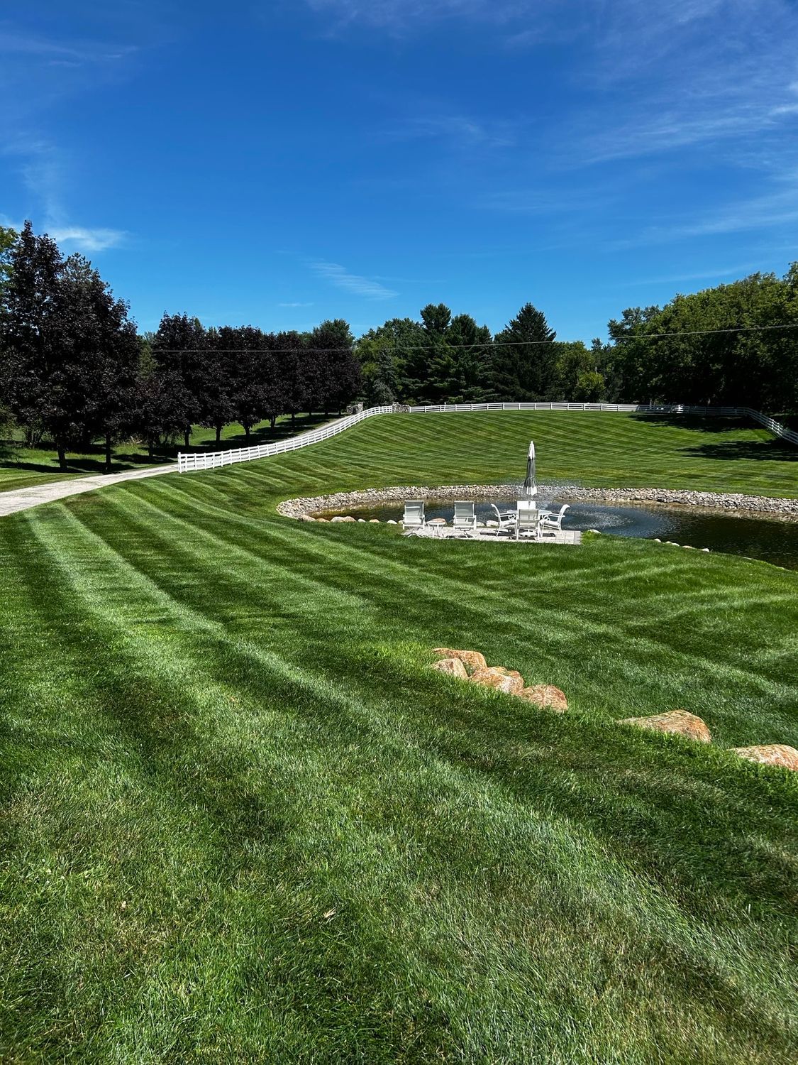 Grassy golf course with rolling fairway, trees, blue sky, and a small water fountain pond in the center