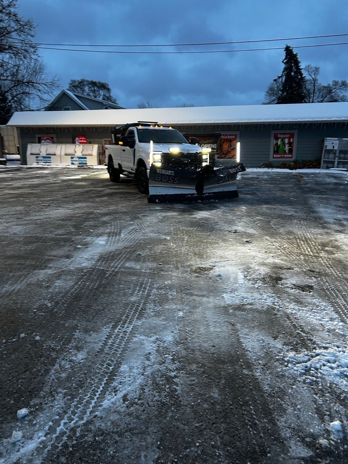 White pickup with plow on icy lot in front of a low building at dusk
