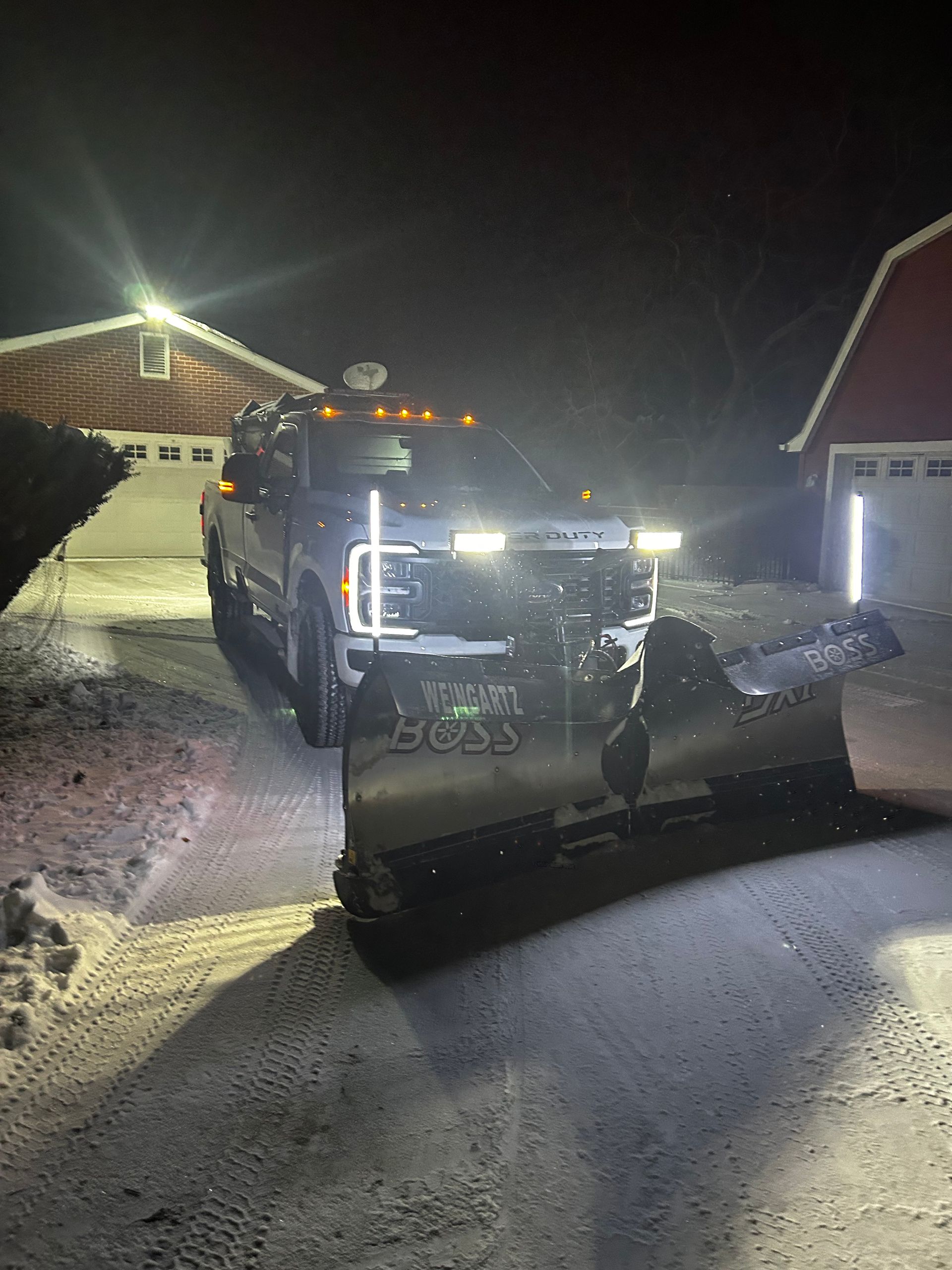 Snowplow clearing a snowy driveway at night between houses, bright headlights shining