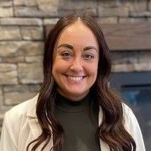 A white woman with long brown hair smiles in front of a stone fireplace