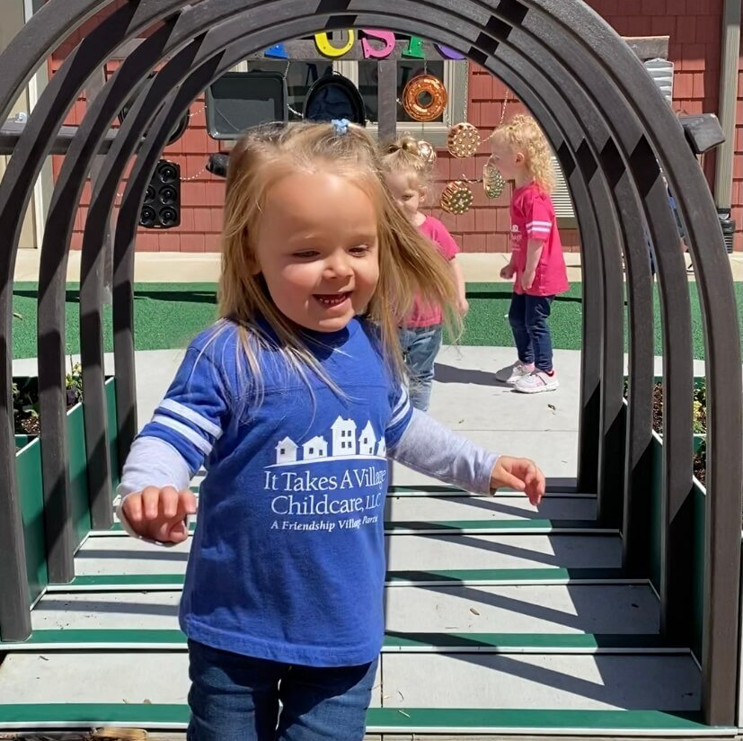 young girl running through a tunnel in the outdoor classroom at It Takes a Village Childcare