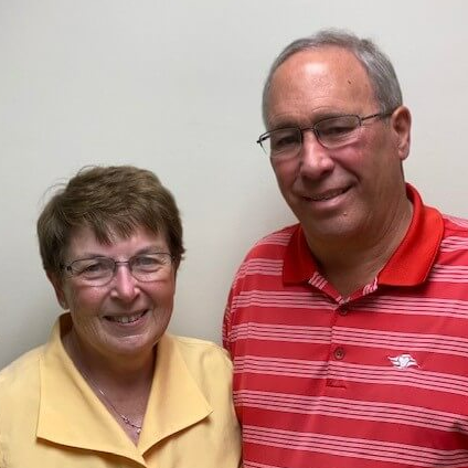 senior couple standing in front of a white wall smiling and posing at the camera for a photo retired couple standing next to each other smiling at the camera