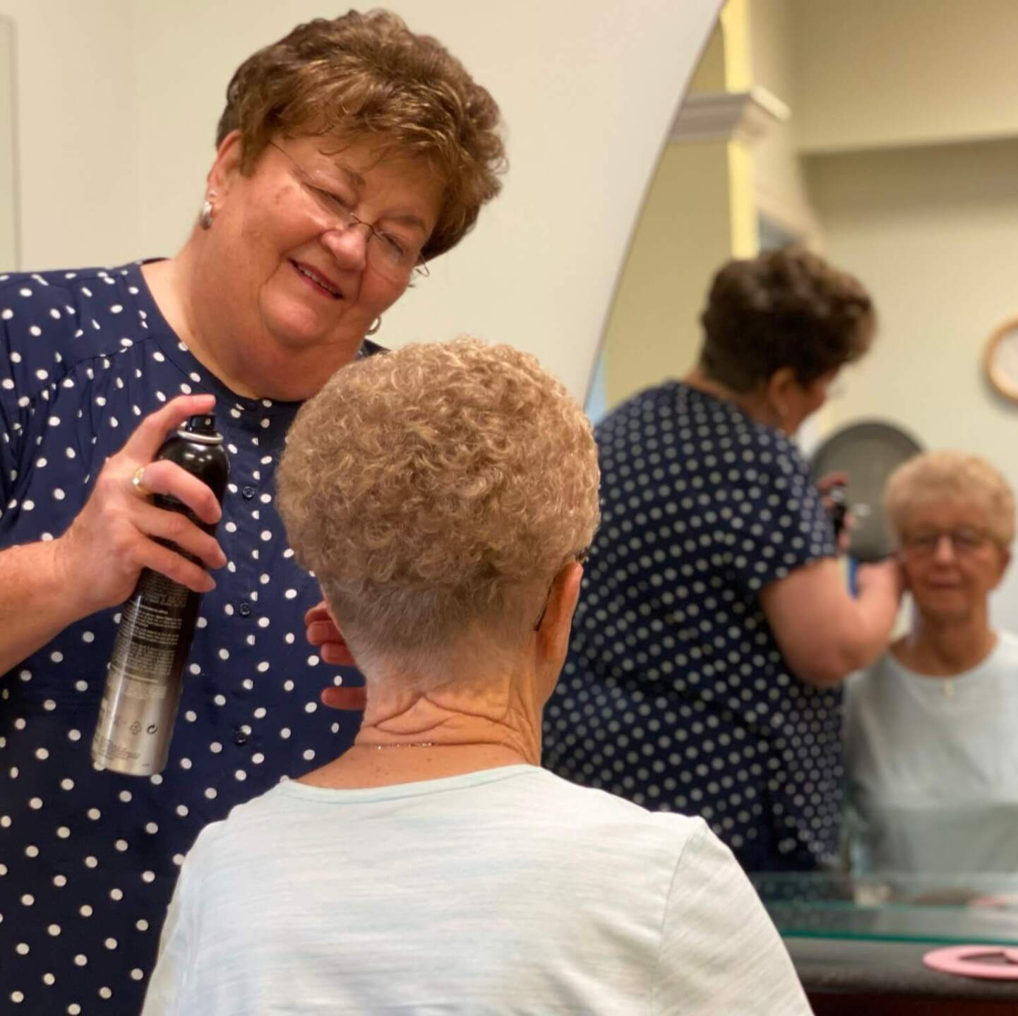 Beautician hair spraying a residents hair while she looks in the mirror in the beauty salon at Landmark Commons