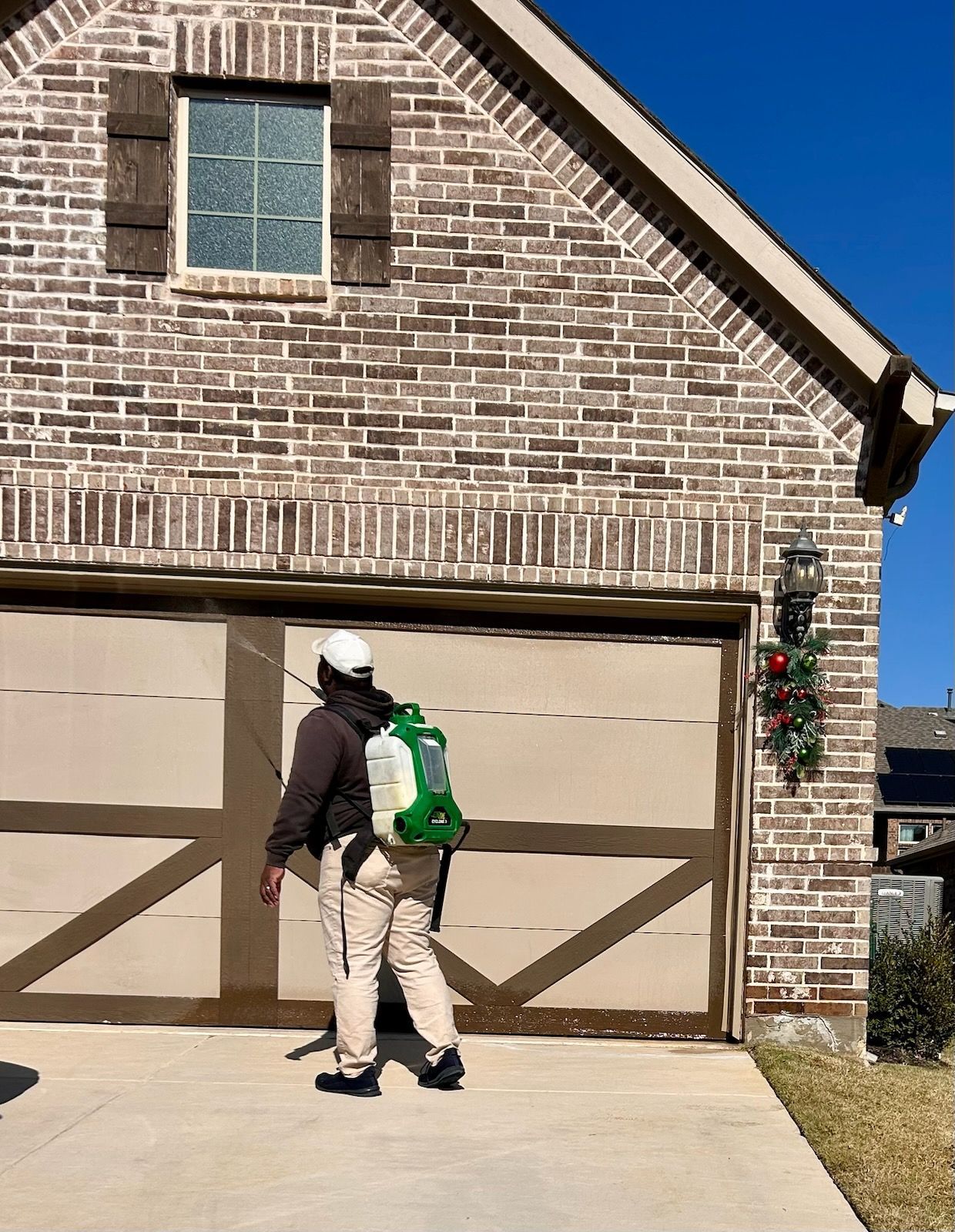 Pest control technician spraying the exterior of a residential garage in North Texas with a backpack sprayer for perimeter pest treatment.