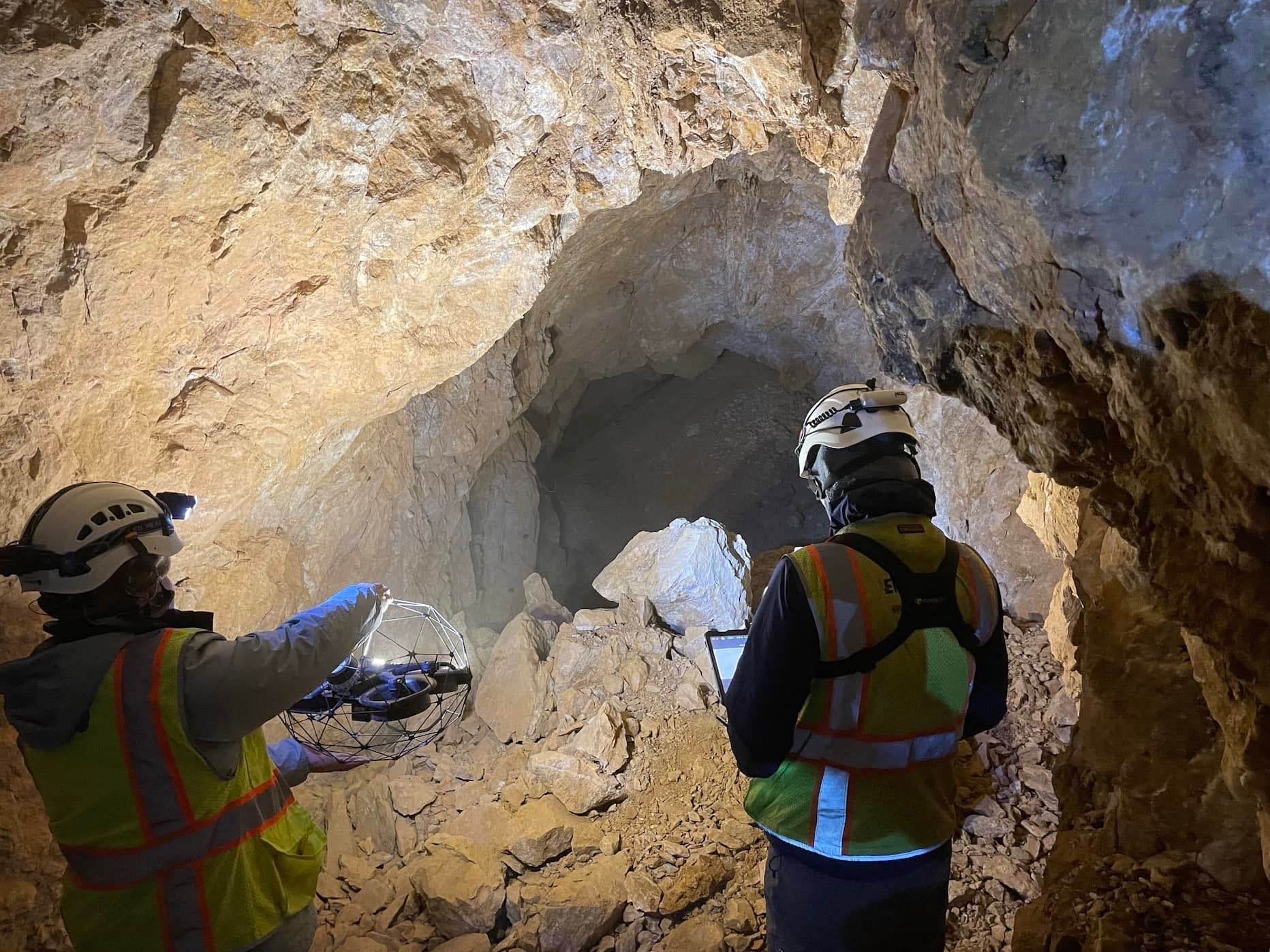 Two workers in hard hats inspect a rocky tunnel with flashlights and safety vests.