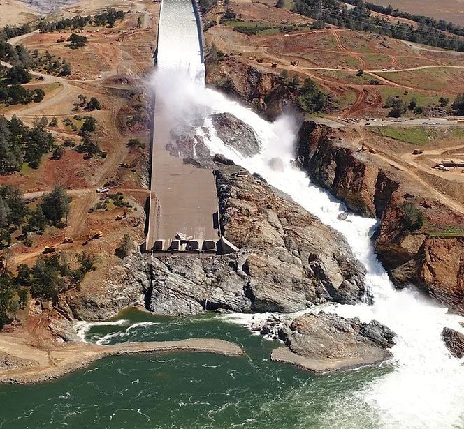 Waterfall surging over a dam spillway into a rocky river gorge, with green water below.
