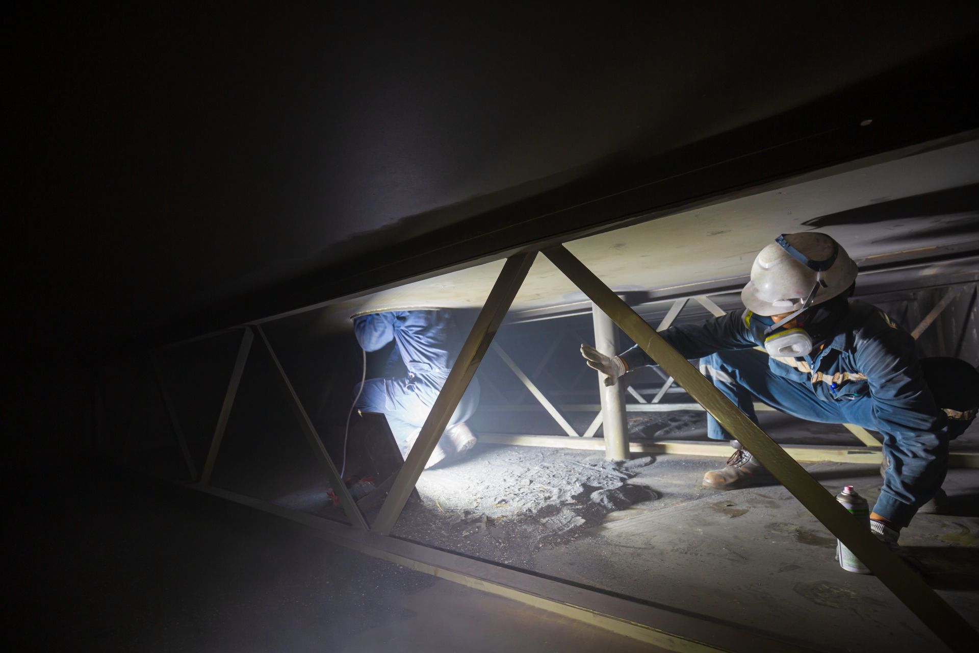 Worker in helmet and blue jacket inspecting a dimly lit metal structure in an industrial space