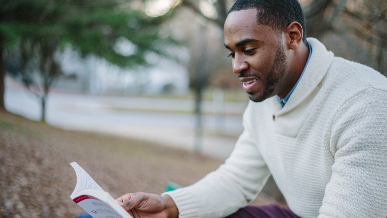 A man in a white sweater is sitting on the ground reading a book.