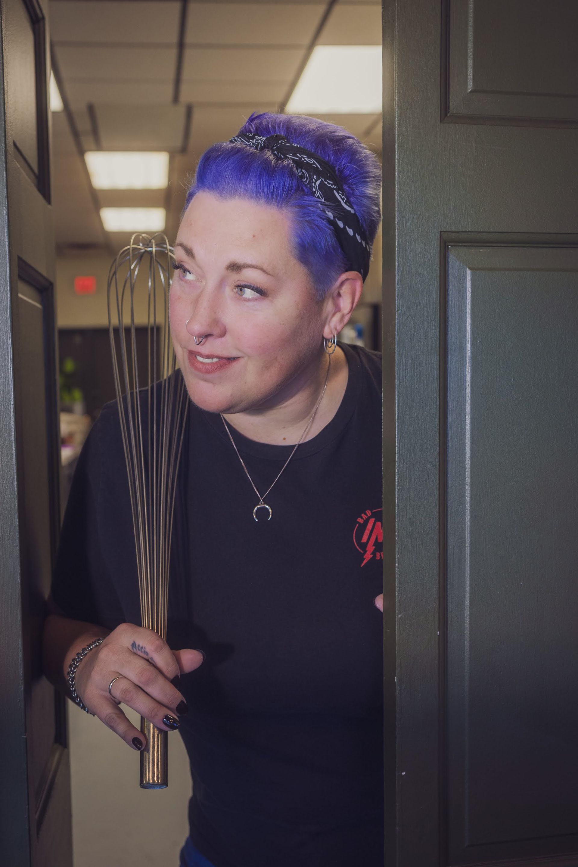 women with purple hair holding large whisk looking out a set of doors