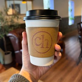 a persons hand holding a hot coffee with a coffee sleeve stamped with ch in a circle
