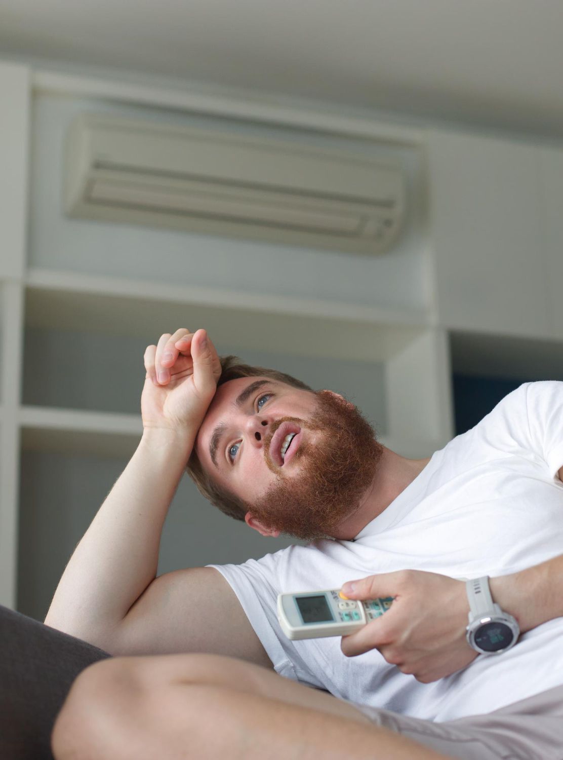 Man reclining on a couch, holding a remote and looking up at an air conditioner.