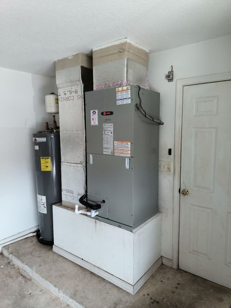 Two gray outdoor HVAC units beside a house on a gravel strip near a brick wall and window.