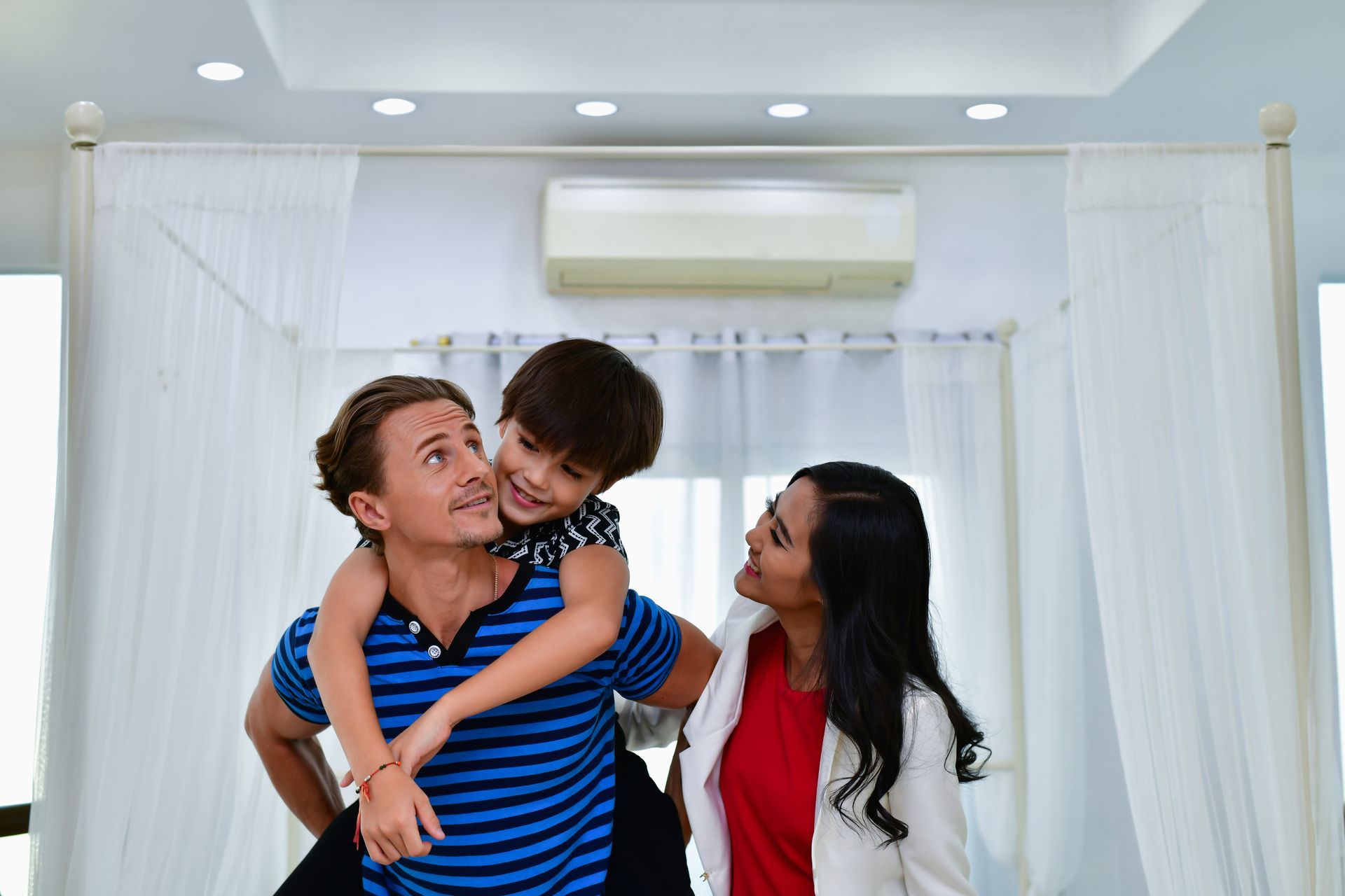Man carrying a smiling child on his back as a woman looks on in a bright room