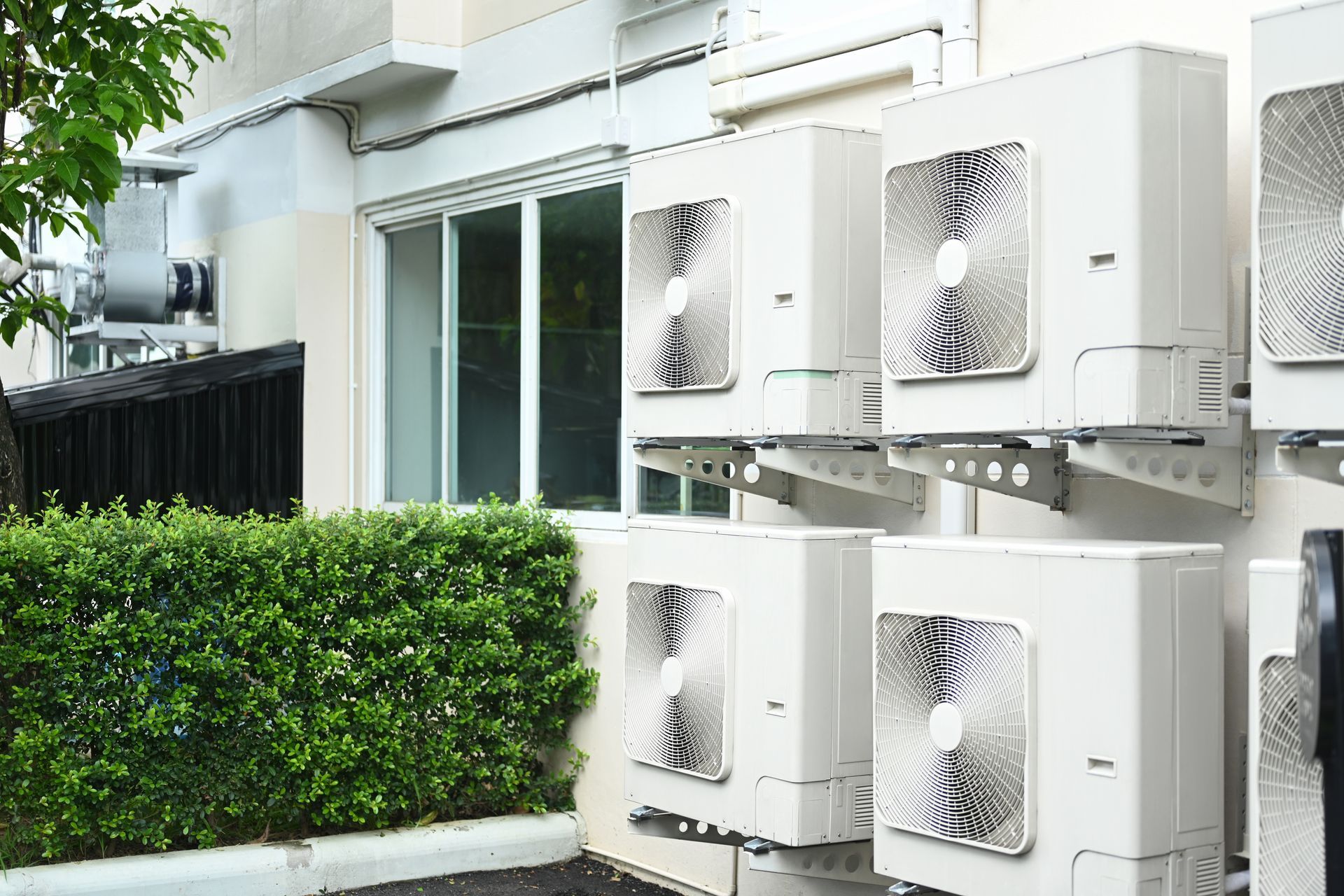 Outdoor wall-mounted air conditioner units beside a modern house and green hedge.