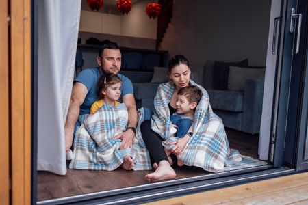 Family sitting on a rug by an open sliding door, wrapped in plaid blankets, with a sofa in the background