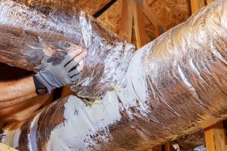 Gloved hand sealing a silver insulated duct in an attic with spray foam insulation