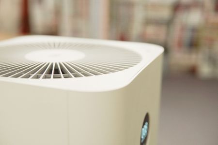 Close-up of a white air purifier with a vented top in a blurred indoor setting