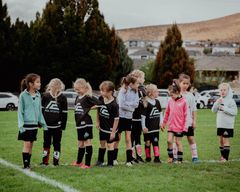Coach kneels with children on a grassy field during an outdoor sports activity, all gathered in a circle