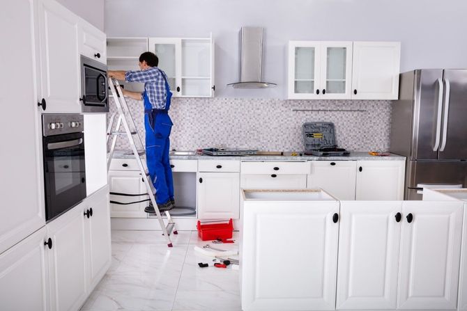 Person on ladder fixing a built-in cabinet in a white kitchen with tools on the floor