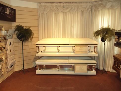 White casket in a funeral home display room with draped curtains and floral arrangements