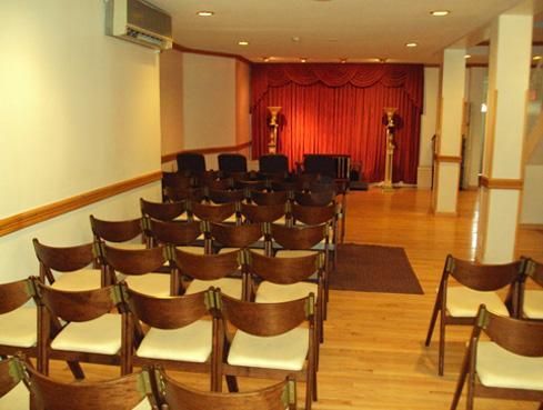Rows of wooden chairs facing a small stage with red curtains in a hall