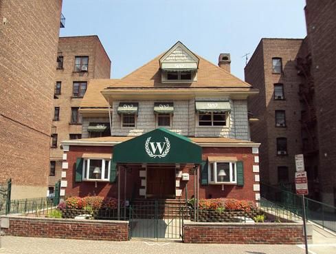 Brick building entrance with green awning and white “W” logo, flanked by taller apartment buildings.
