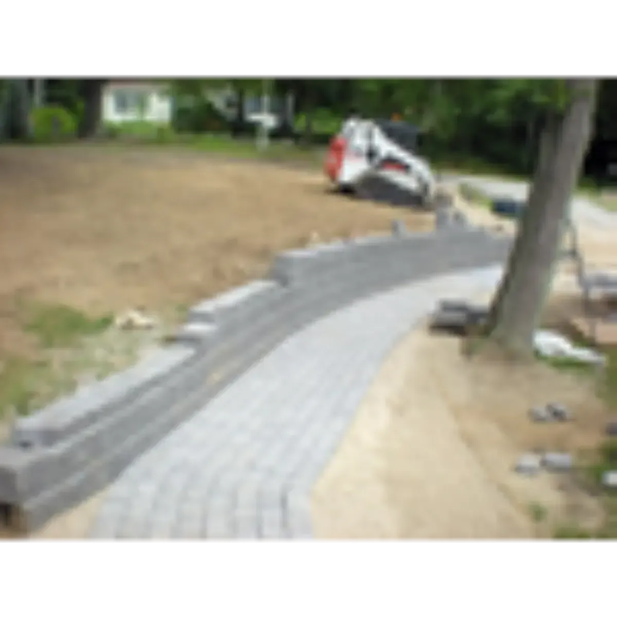 Curved gray brick walkway under construction beside a dirt yard and tree trunk