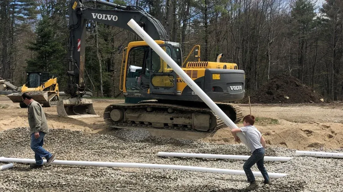 A boy is holding a pipe in front of a volvo excavator.