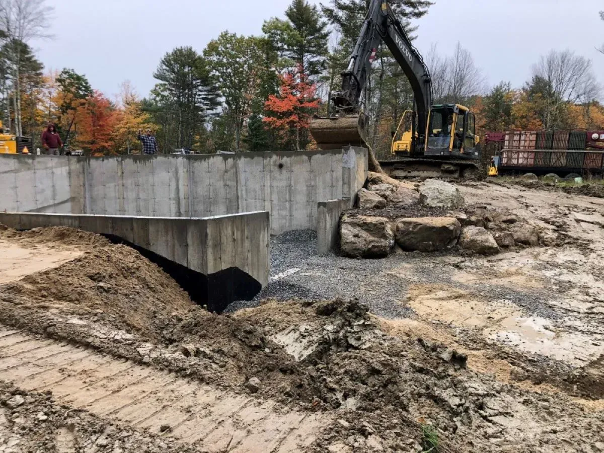 A large excavator is digging a hole in the ground in front of a concrete wall.