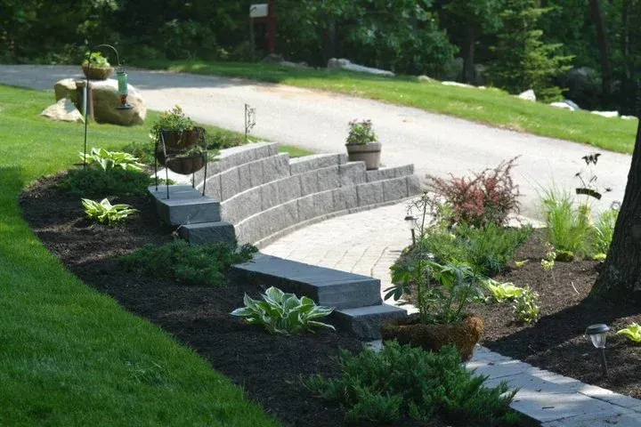 A stone walkway leading to a driveway surrounded by plants and bushes