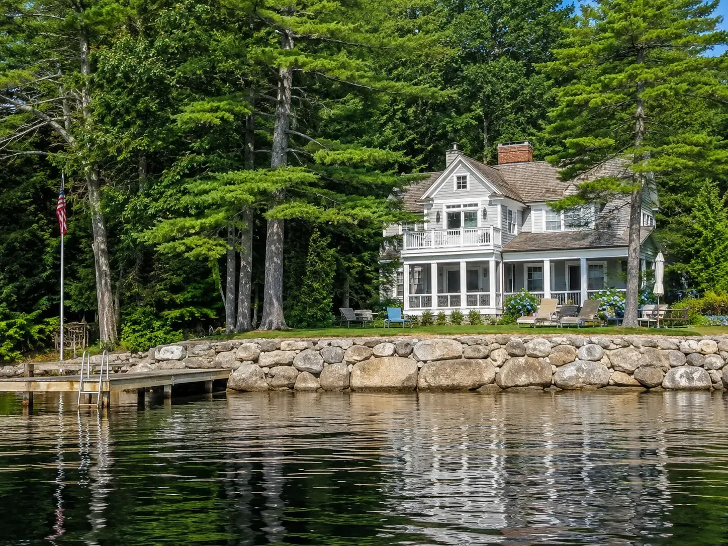 White lakeside house with a stone wall, dock, and tall green trees reflected in calm water