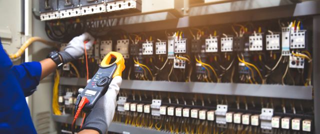 Techniker prüft in einer Fabrikumgebung einen elektrischen Schaltschrank mit einem Multimeter und Werkzeug.