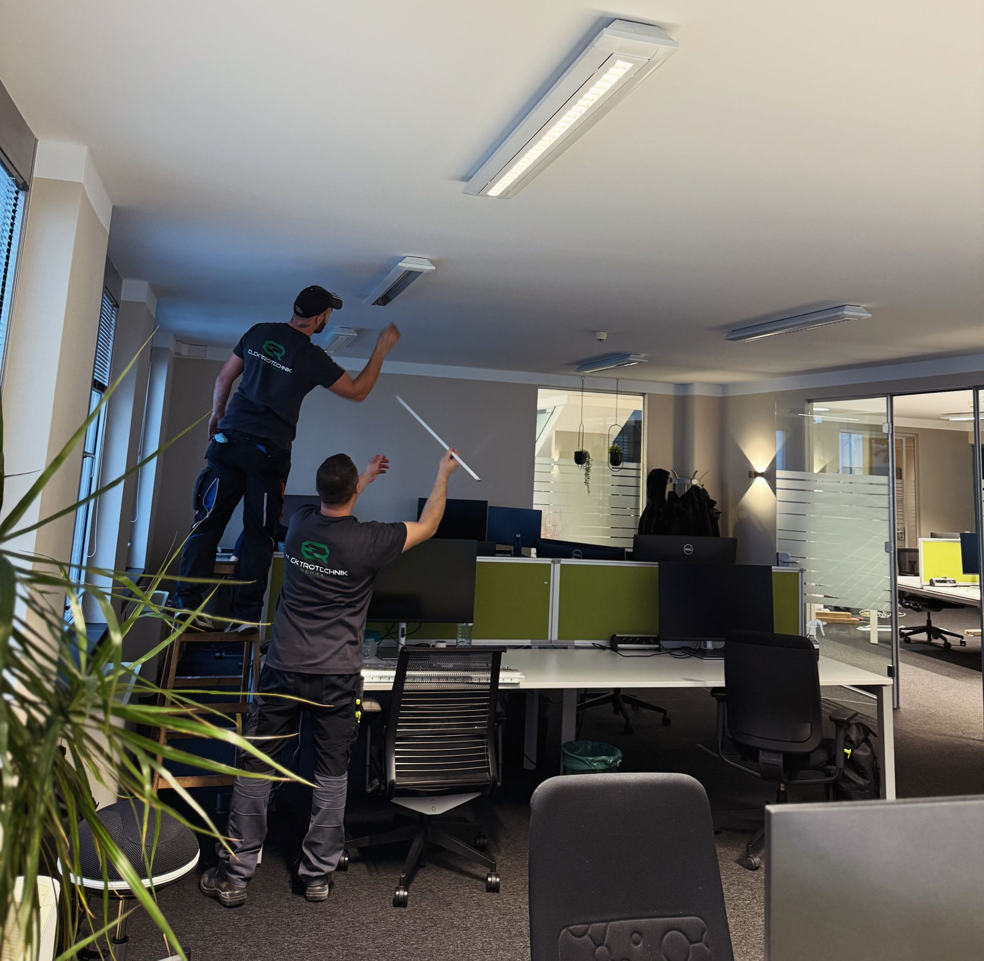 Zwei Personen tauschen Deckenleuchten in einem Büro mit Schreibtischen und Computern aus.