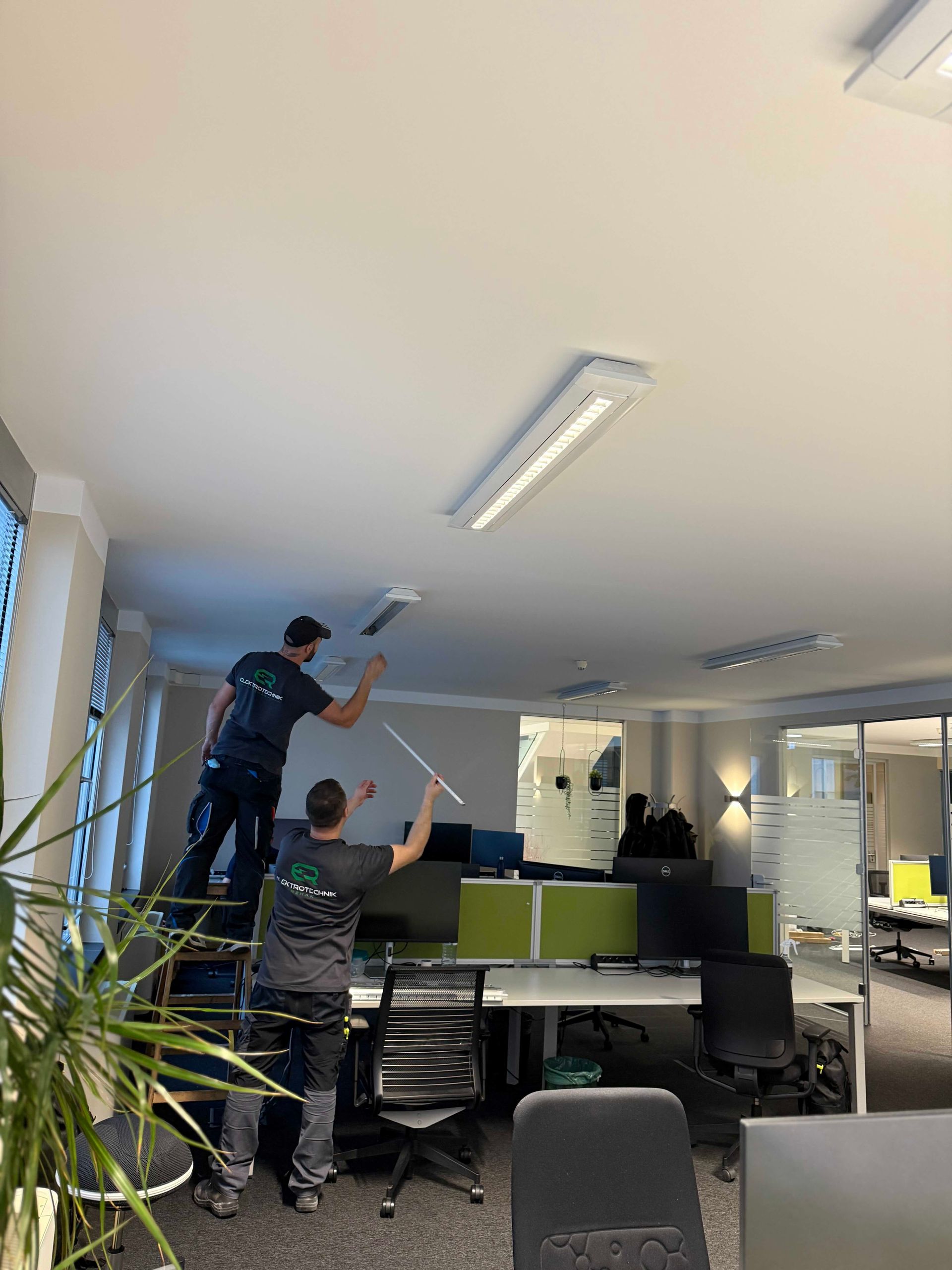 Zwei Personen streichen die weiße Decke über Schreibtischen und Stühlen in einem modernen Büro.