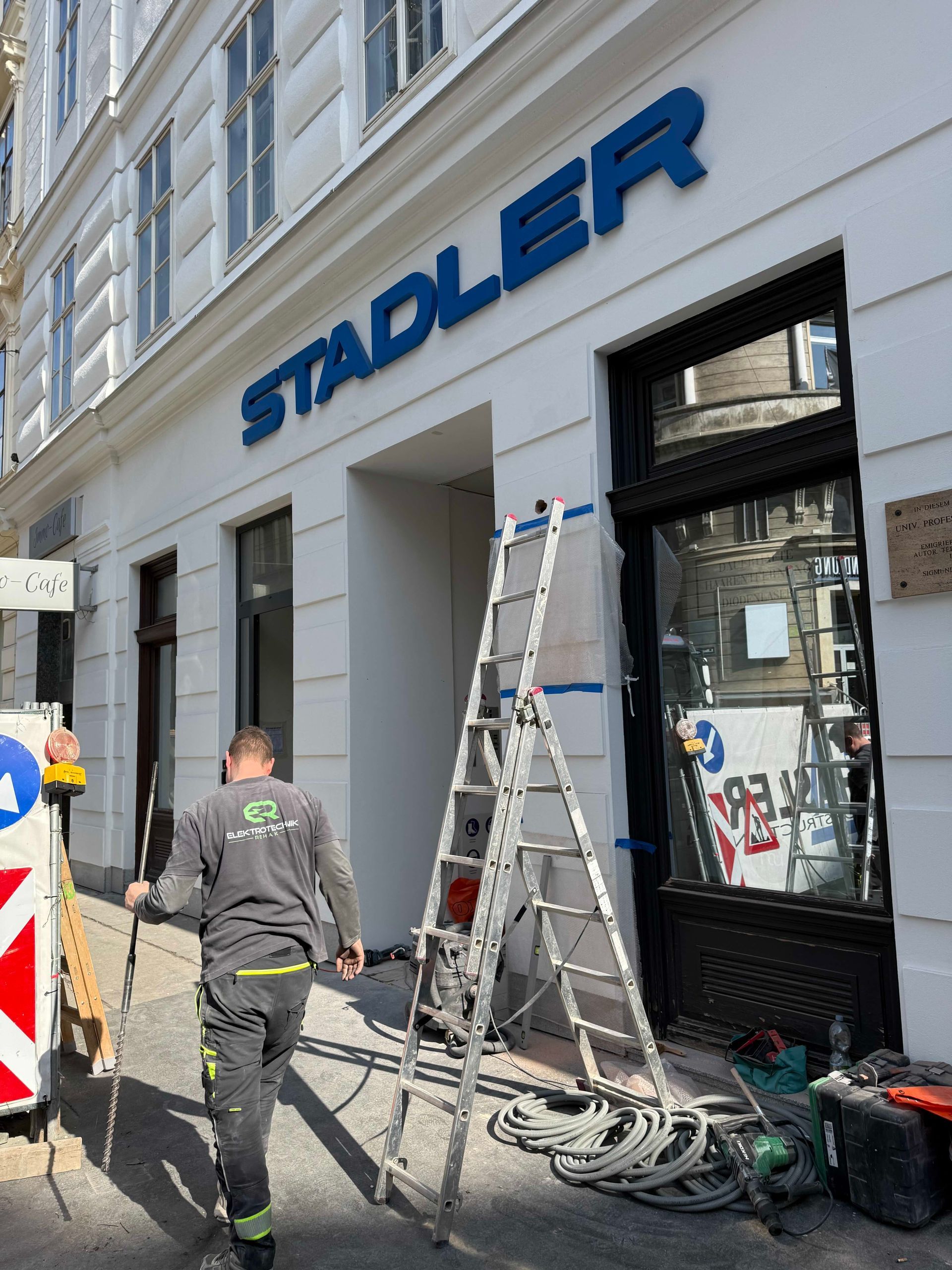Arbeiter vor einem STADLER-Ladenlokal, Leitern, Werkzeuge und Kabel auf dem Bürgersteig.