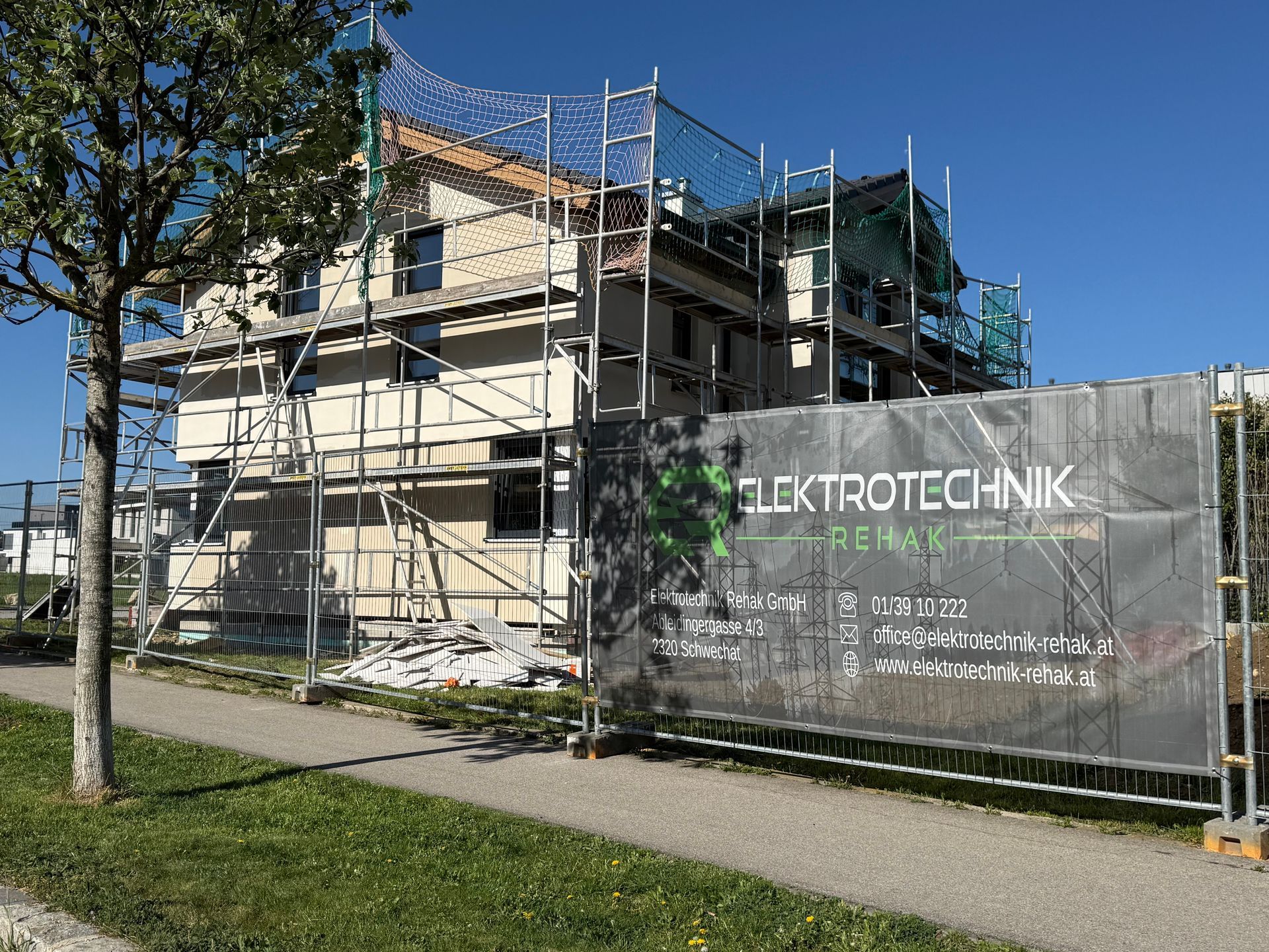 Ein im Bau befindliches Gerüst neben einem Zaunschild mit der Aufschrift „ELEKTROTEHNIK“ an einem sonnigen Tag