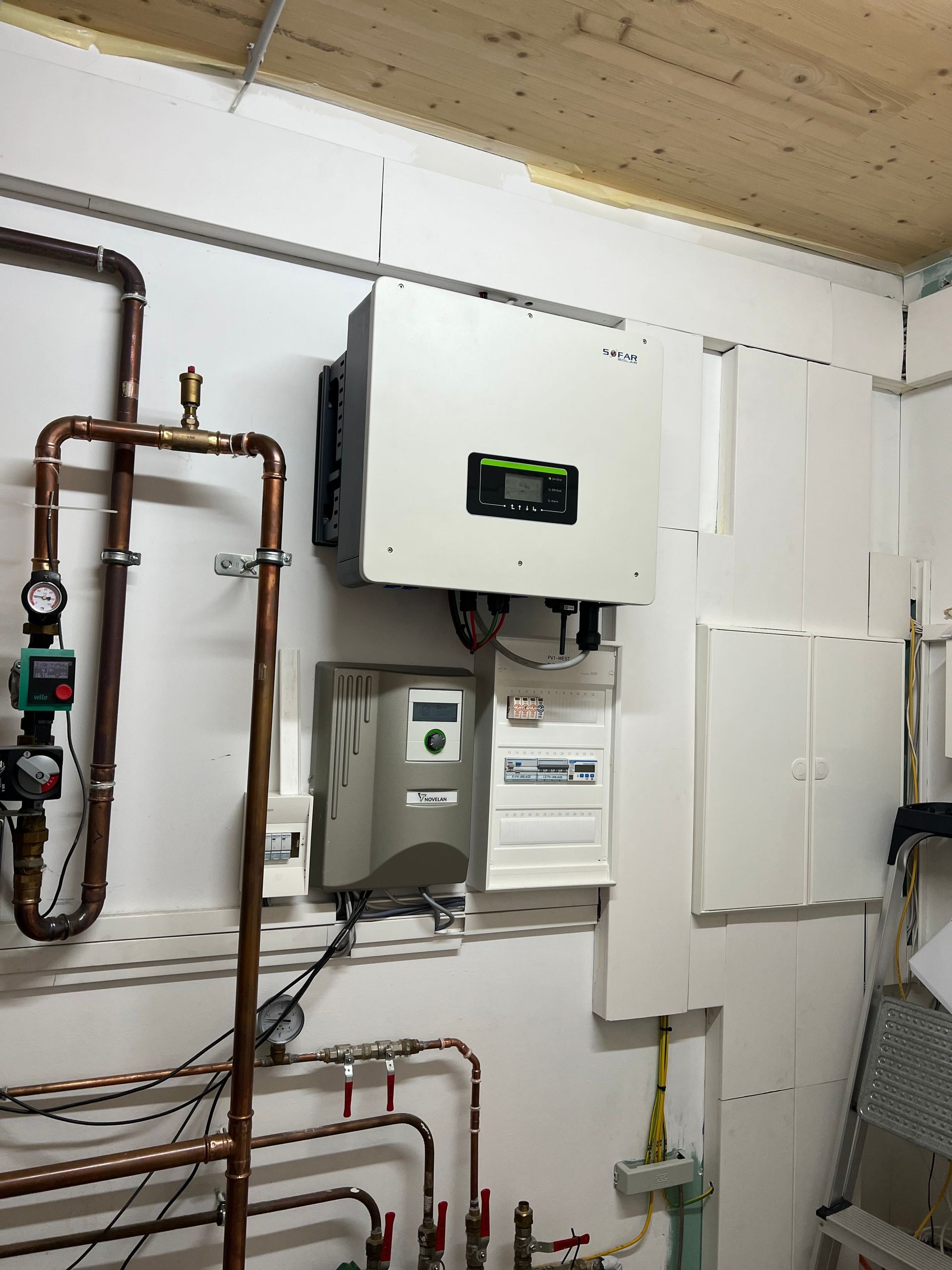 Hauswirtschaftsraum mit wandmontiertem Heizkessel und Rohrleitungen sowie elektrischen Schaltkästen an einer weißen Wand.