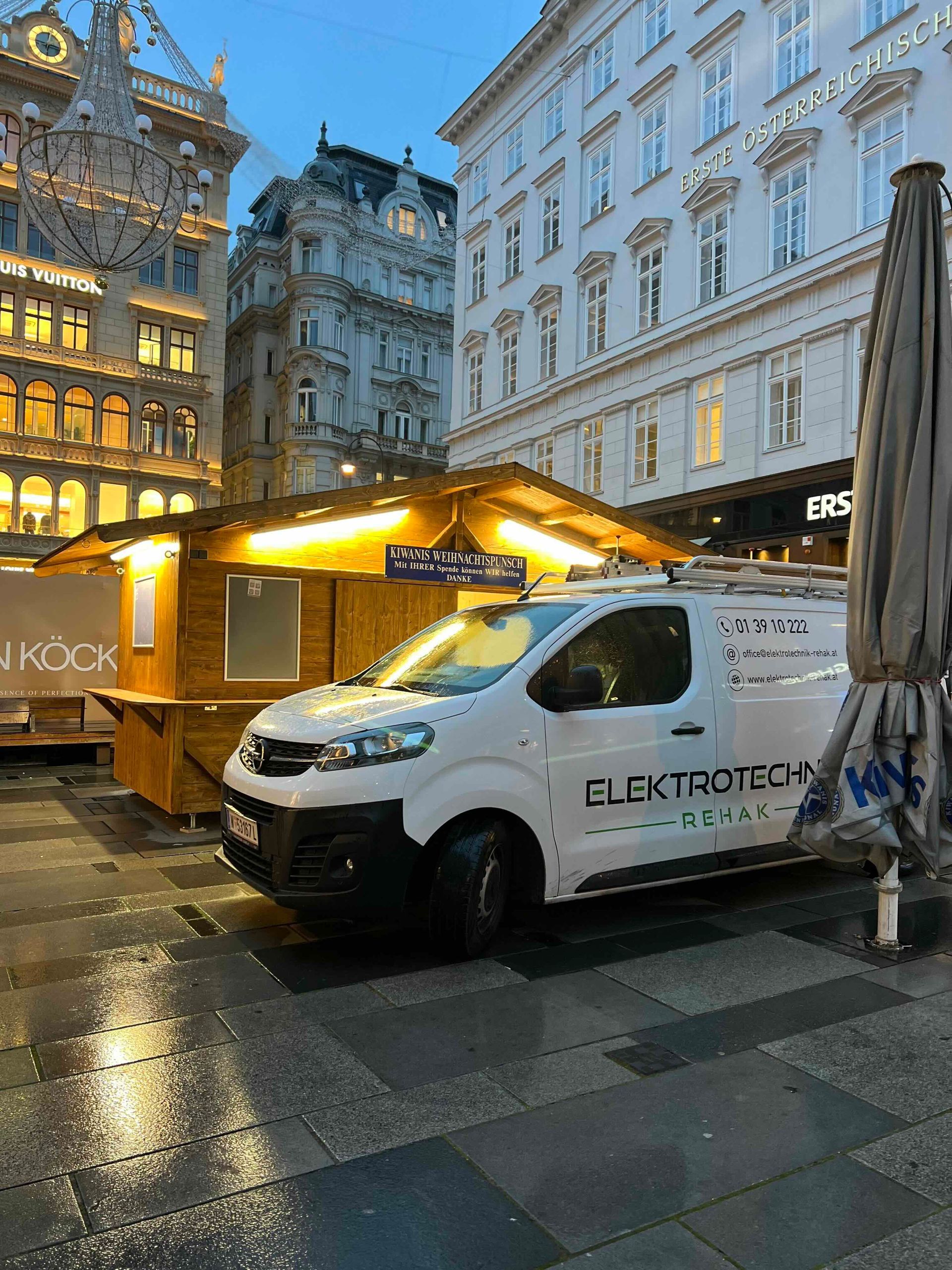 Ein weißer Lieferwagen parkt in der Abenddämmerung neben einem beleuchteten Marktstand auf einer nassen Straße.
